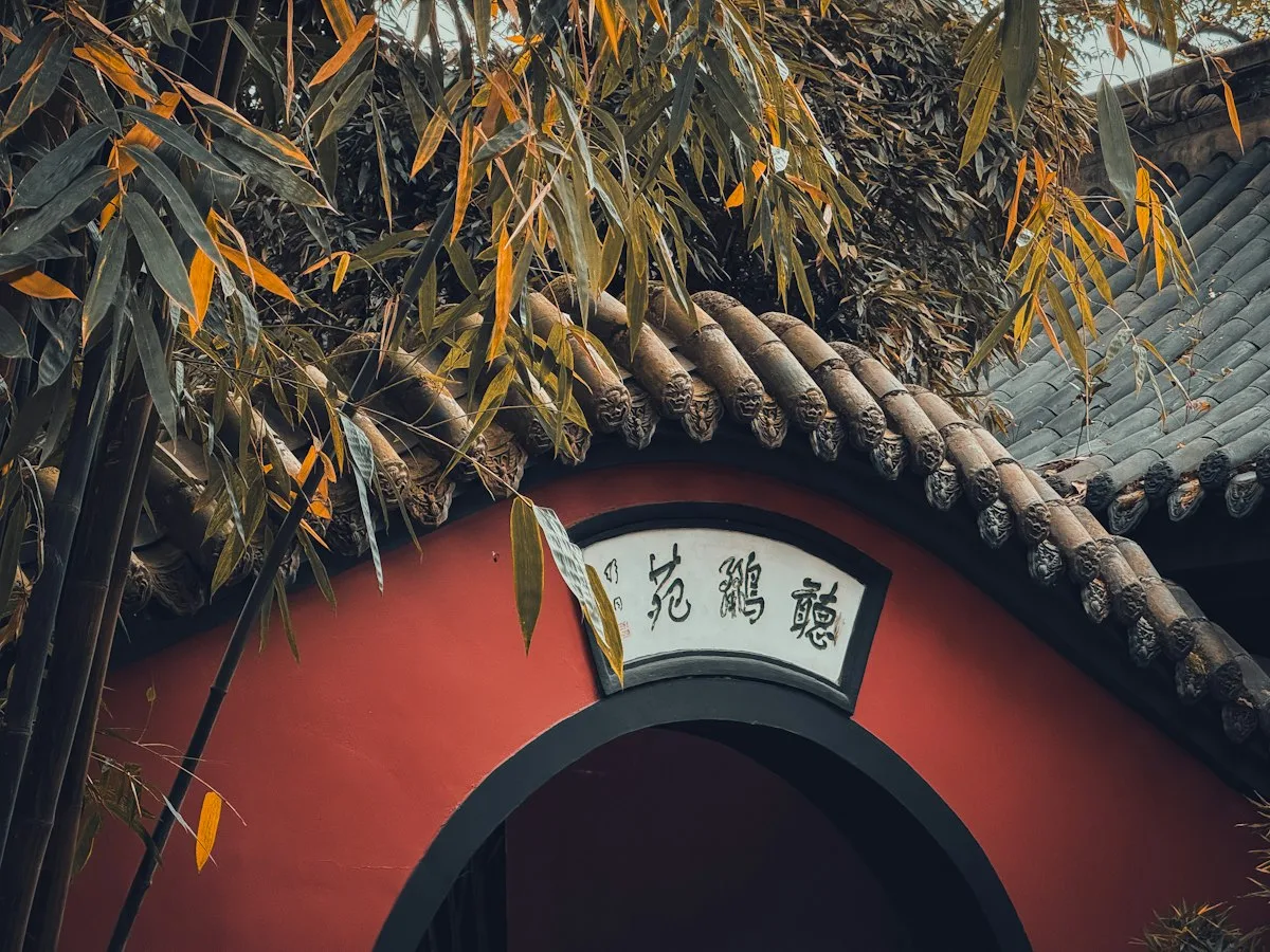 Um muro de jardim vermelho com telhado de telhas cinzentas e um arco sob uma placa branca de caligrafia chinesa, emoldurado por bambu