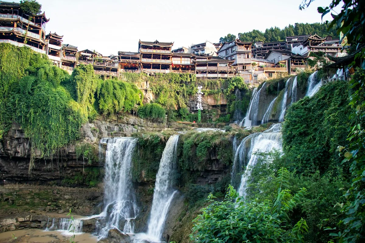 The two-tier waterfall at Furong Town dropping past a cliff of green foliage, with wooden stilt houses and tiled roofs along the top of the gorge