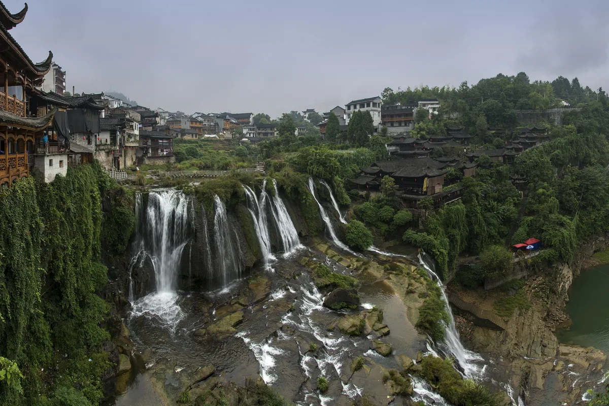The two-tier waterfall at Furong Town dropping through the gorge, with Tujia stilt houses on the left and the old town built along the cliff above