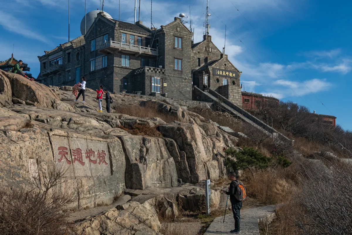 The stone weather station and observatory buildings on the summit ridge of Mount Tai, with the red rock inscription Badi Tongtian below and a hiker on the stone path