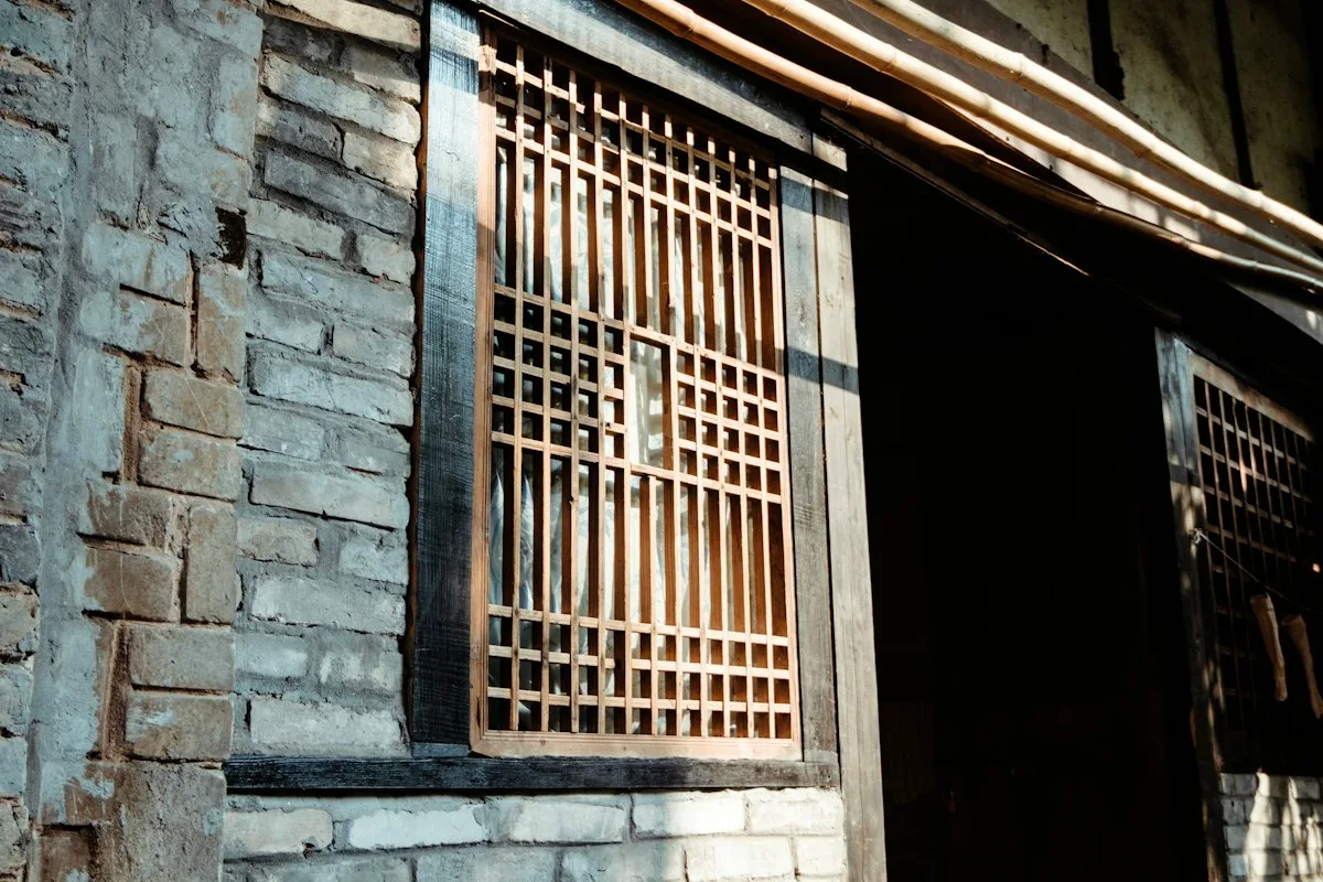 A wooden lattice window set into the old brick wall of a traditional guesthouse, sunlight falling across it