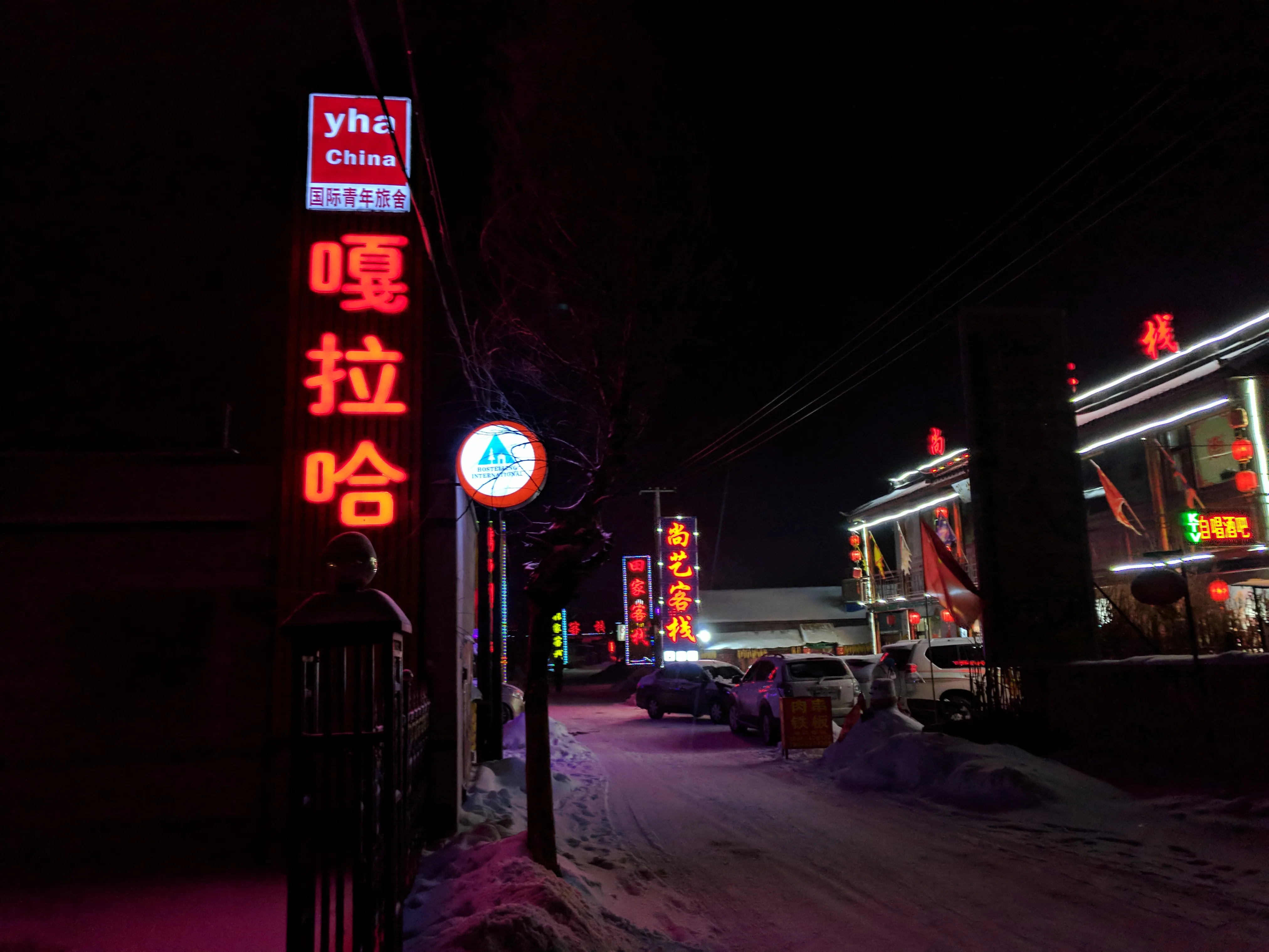 A youth hostel with a red neon sign on a snowy street in Jilin at night, Chinese shopfronts and parked cars alongside (Photo: Enming Yan via Wikimedia Commons)