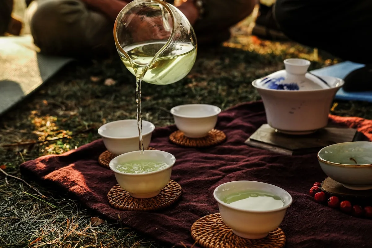 Green tea being poured from a glass pitcher into small cups on a bamboo tray
