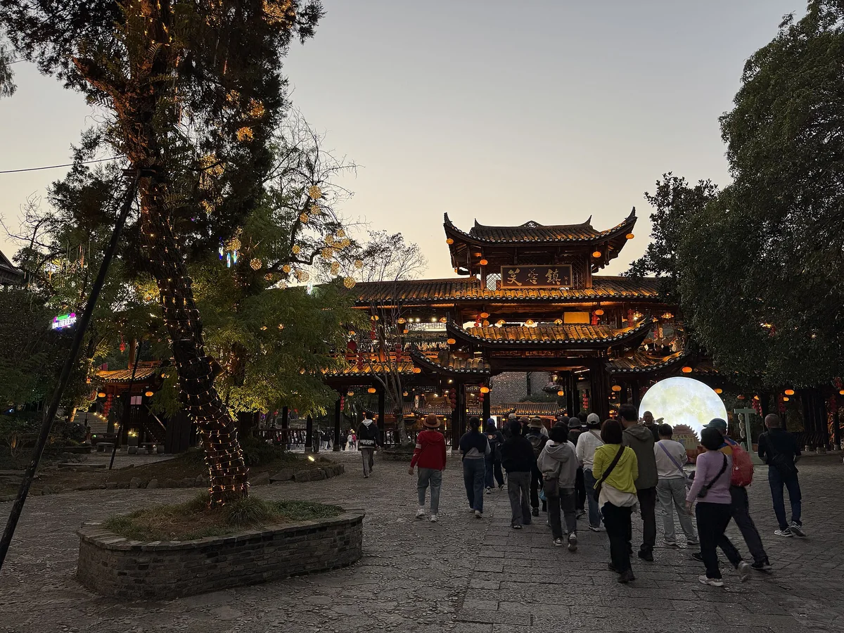 The entrance gate of Furong Town at dusk, with lights on the timber gatehouse and a tour group filing through the stone plaza (Photo: Codas via Wikimedia Commons)