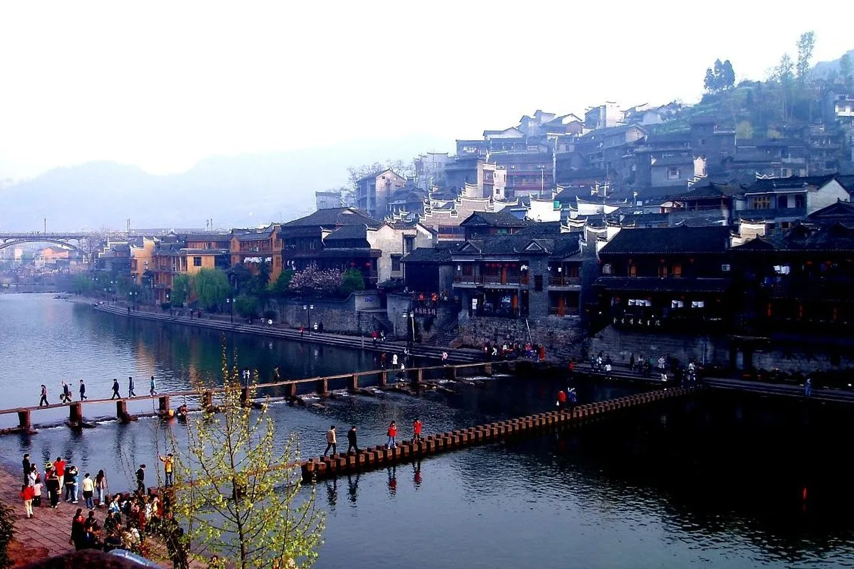 The Tuo River at Fenghuang, with people crossing the jumping stones in the foreground and stilt houses stacked along the far bank below the town (Photo: Yu Hui via Wikimedia Commons)