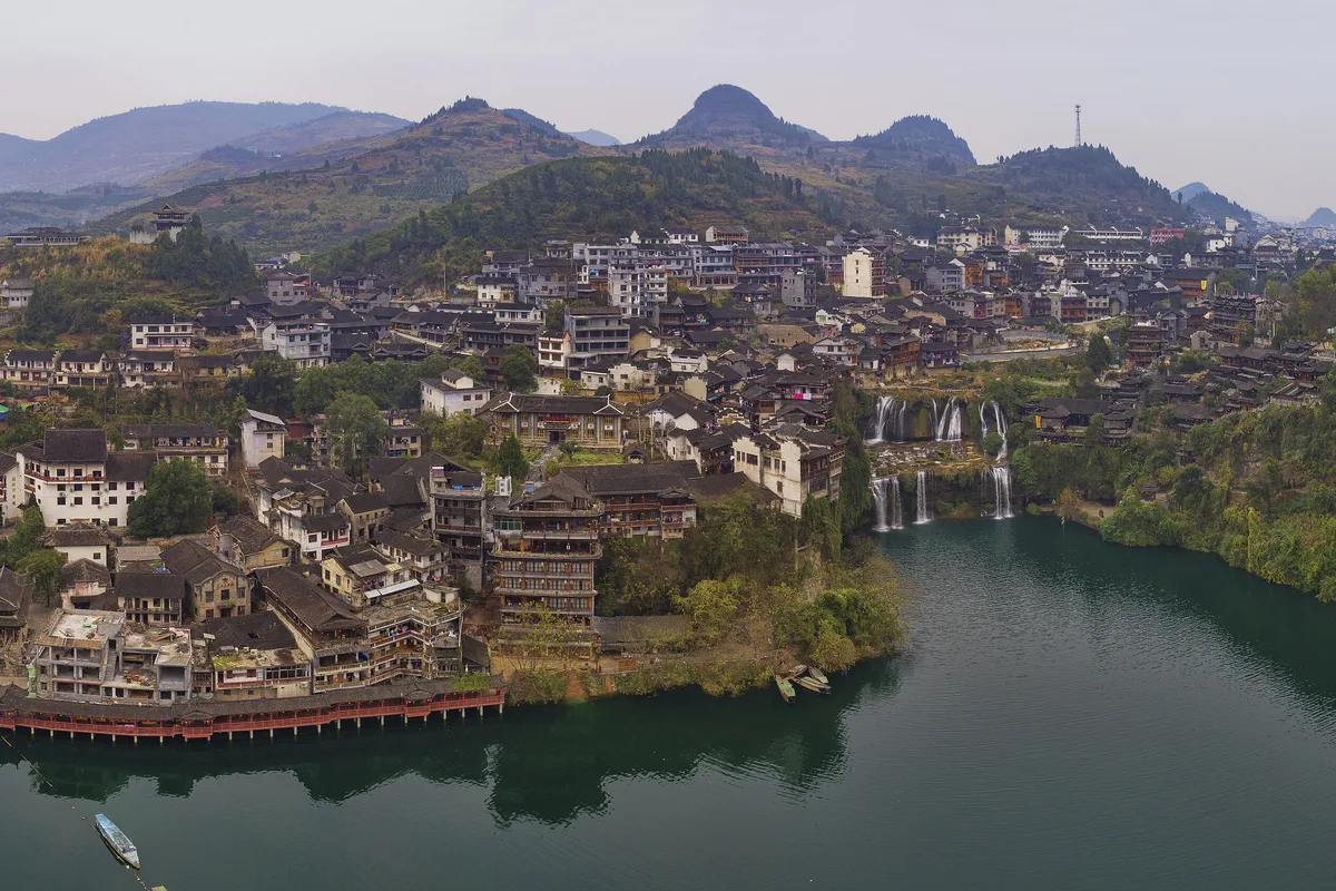 An aerial view of Furong Town seen from above the river bend, with the waterfall dropping into the water on the right, boats moored along the bank and the whole town visible in a single frame (Photo: Chensiyuan via Wikimedia Commons)