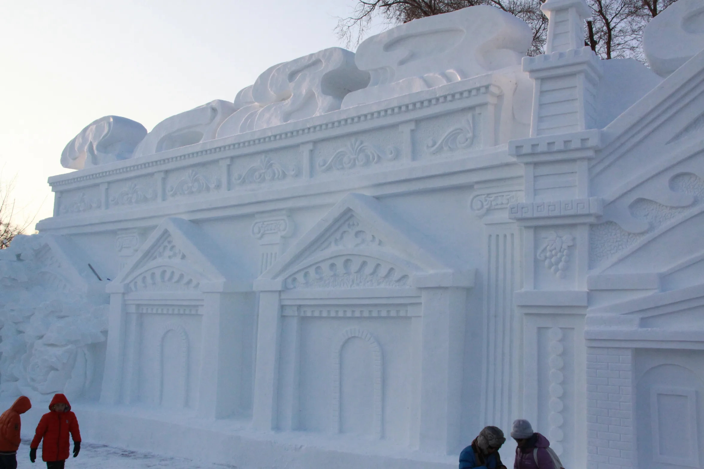 Visitantes passando por uma escultura de neve finamente entalhada de um prédio clássico com colunas e relevos, na Exposição de Esculturas de Neve da Ilha do Sol em Harbin (Photo: Caliva via Wikimedia Commons)