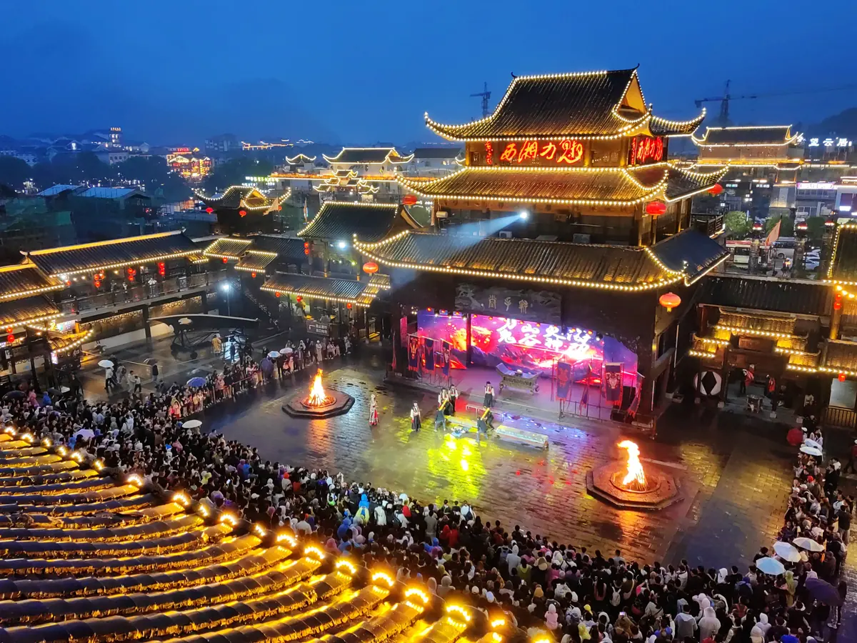 Crowds gathered around bonfires in the courtyard of the Charming Xiangxi theatre at night, in front of the illuminated outdoor stage