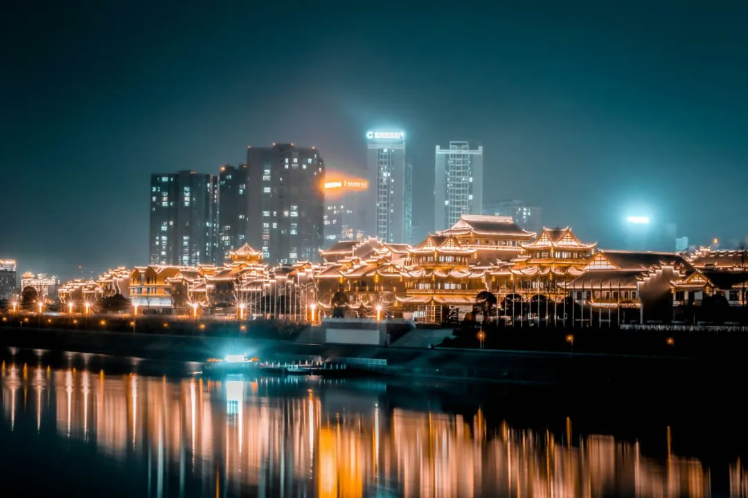 The Dayong Ancient City complex in Zhangjiajie lit up at night, its tiered roofs reflected in the river below the modern city skyline
