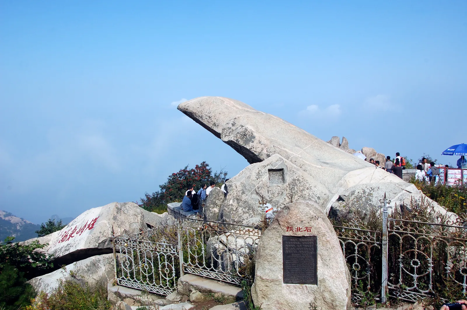 The cantilevered Gongbei Stone on the summit ridge of Mount Tai behind a low iron fence and a stone plaque, with visitors gathered at the railing beyond (Photo: kanegen via Wikimedia Commons)