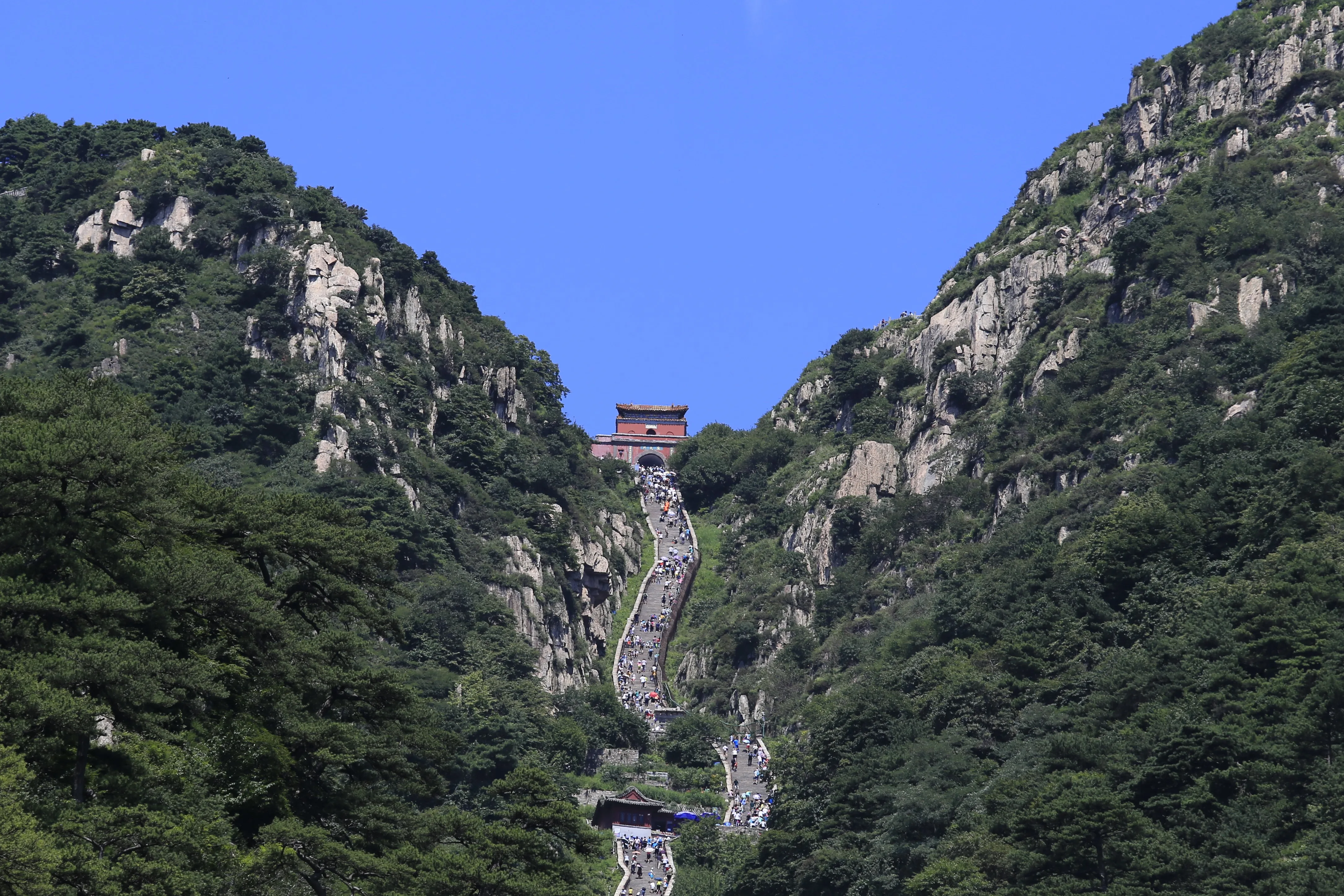 Climbers strung out along the steep stone staircase of the Eighteen Bends on Mount Tai, with the red gatehouse of Nantianmen at the top of the ravine (Photo: Zhangzhugang via Wikimedia Commons)