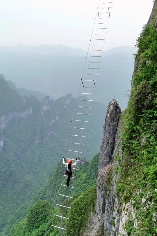 A climber in a helmet partway up the Lingyun Sky Ladder, with the forested valley far below
