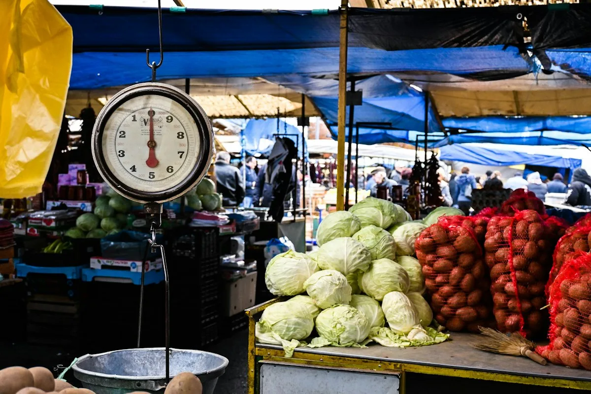 A market stall with a mechanical scale in front, cabbages and netted onions behind
