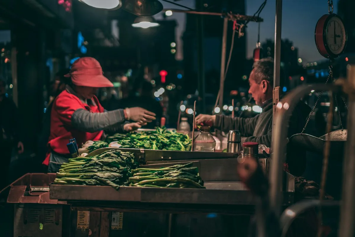 A vendor and customer at a street vegetable stall at night, with a mechanical scale hanging above