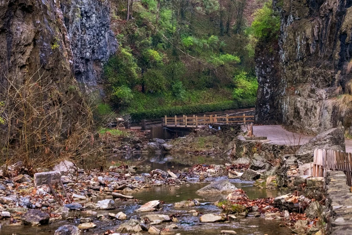 A paved walkway and wooden footbridge crossing a rocky stream between high rock walls, with autumn leaves along the water
