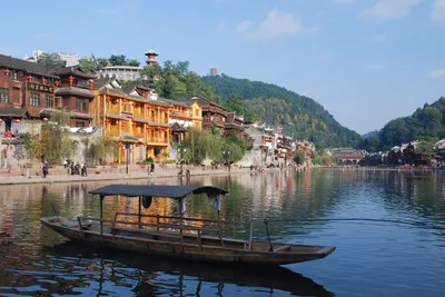 A wooden boat moored on the Tuojiang River at Fenghuang, with stilt houses and red lanterns along the far bank and forested hills behind